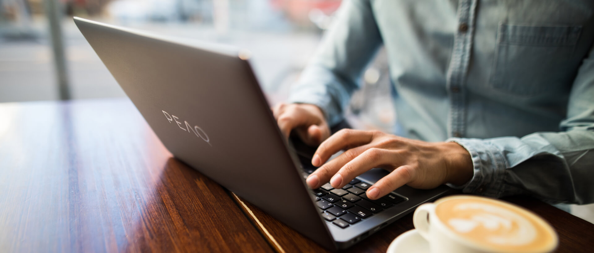 Fiatal férfi ír egy PEAQ laptopon, mellette tele kávéscsésze, egy kávézóban ül, közeli felvétel, panoráma
