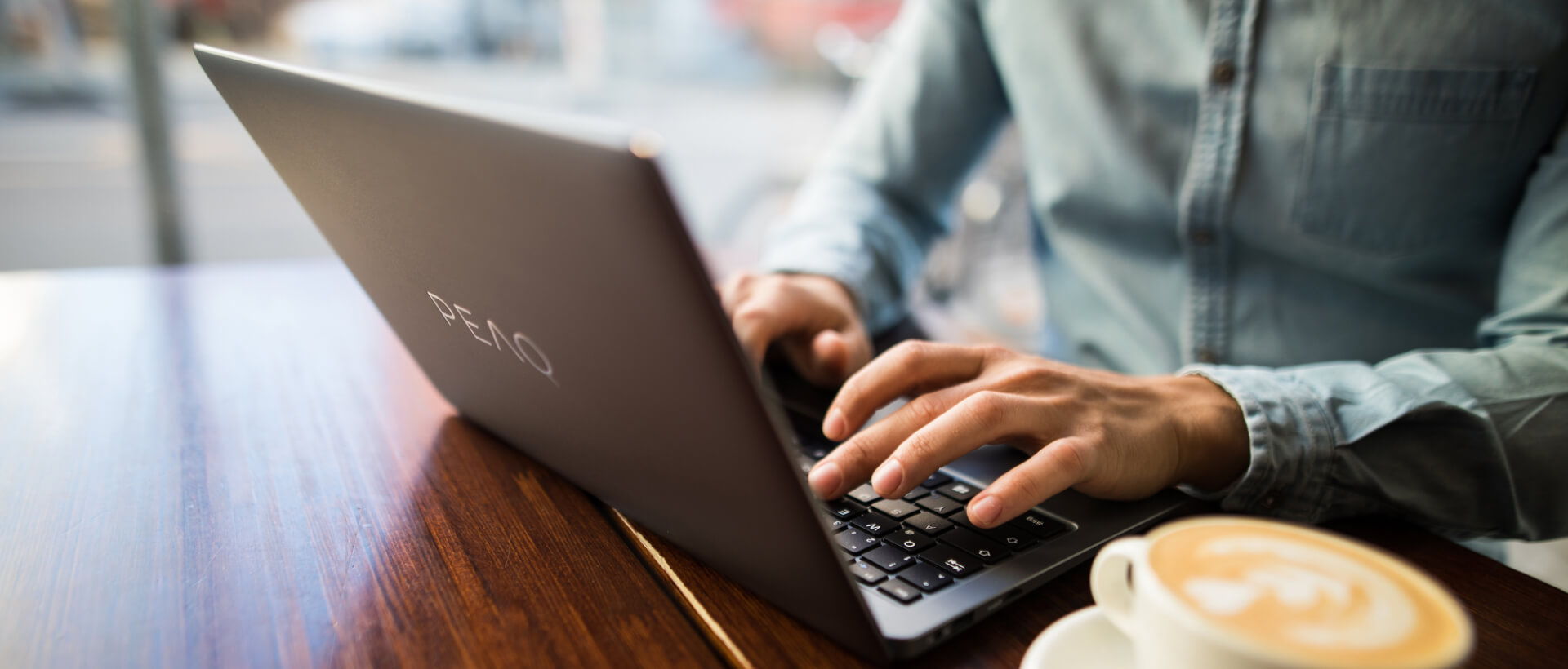 A young man typing on a PEAQ laptop, next to a full coffee cup, sitting in a café, close-up, panorama