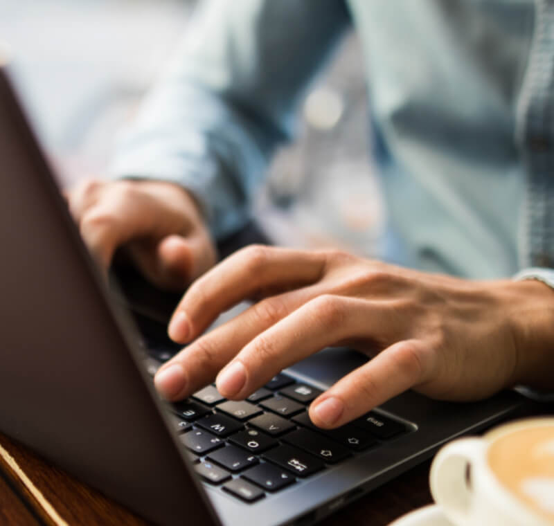 Homem jovem a escrever num portátil da PEAQ, ao lado uma chávena de café cheia, sentado num café, vista de perto