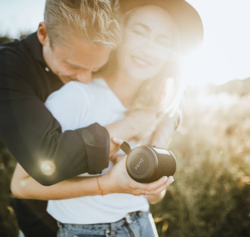 Een jonge vrouw houdt een bluetooth-luidspreker van het merk PEAQ in haar hand, een jonge man omarmt haar van achteren, ze lachen allebei, buiten, zon op de achtergrond