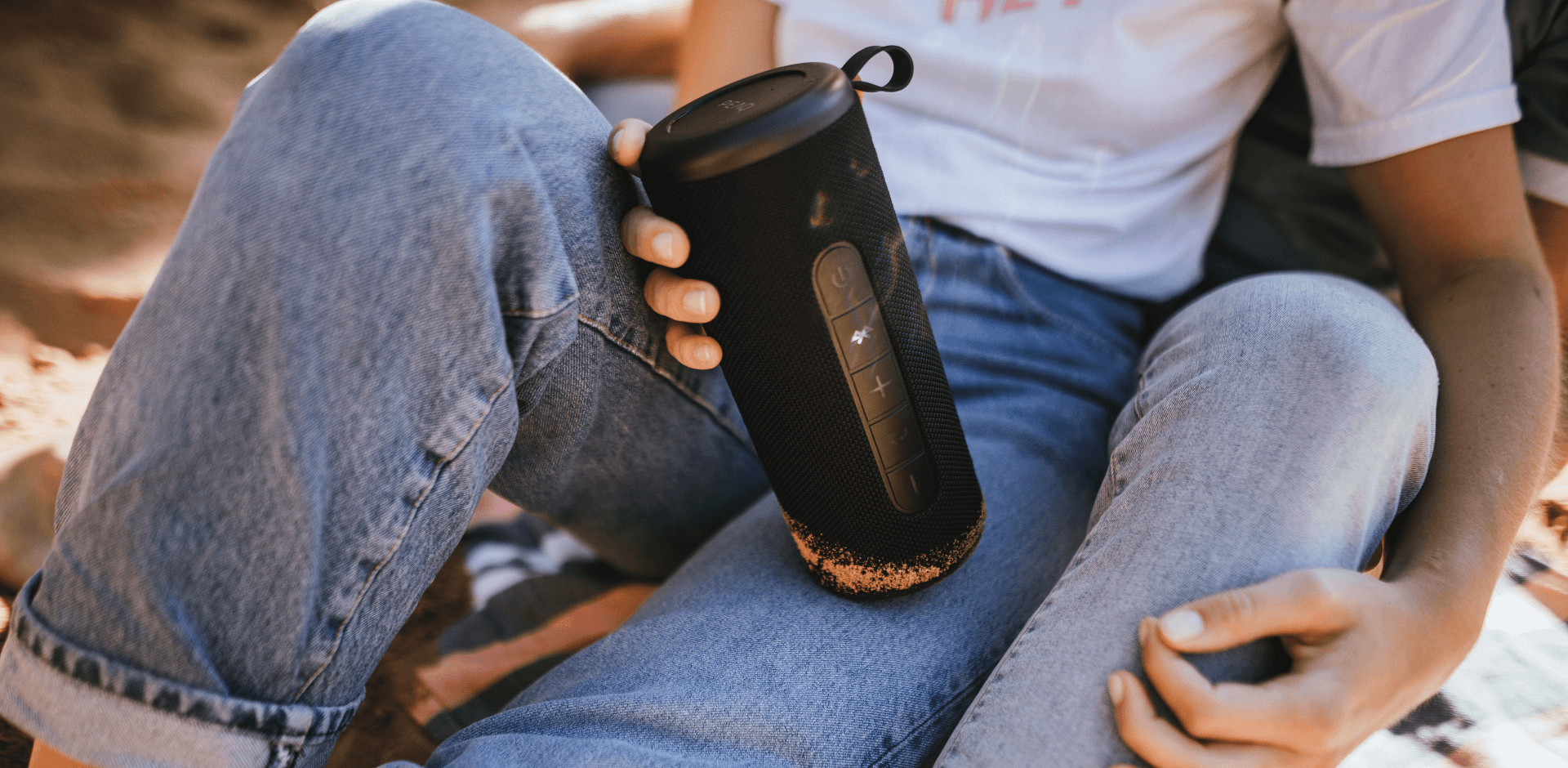 A young woman in jeans holding PEAQ Bluetooth speakers, outdoors, sitting on the beach with her partner, close-up