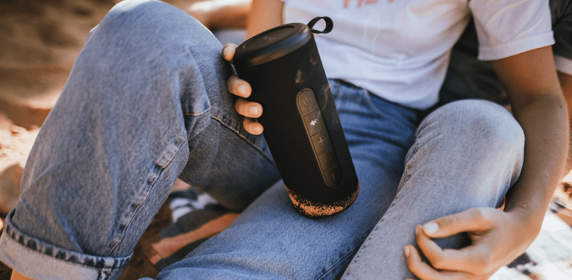 Junge Frau mit Jeanshose hält einen Bluetooth Lautsprecher von PEAQ in der Hand, im Freien, sitzt am Strand mit ihrem Partner, Nahansicht