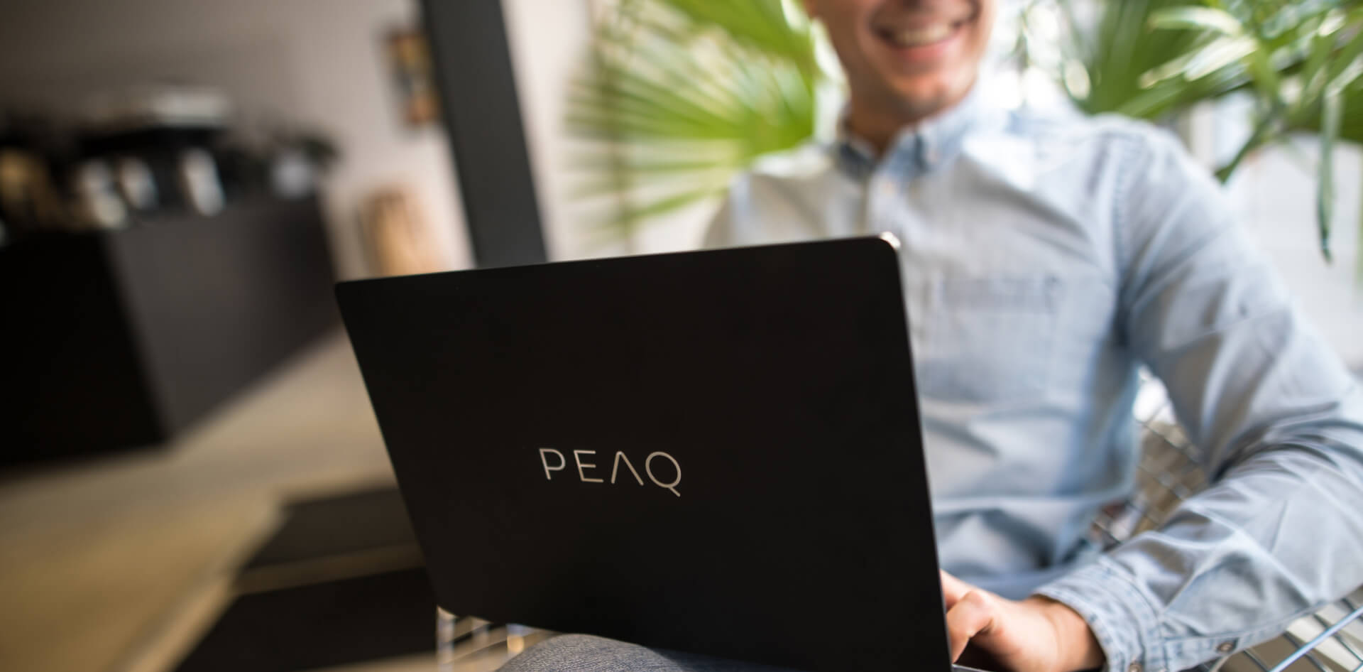 Homem jovem com portátil da PEAQ no colo, sorridente, sentado no escritório ou café, vista de perto