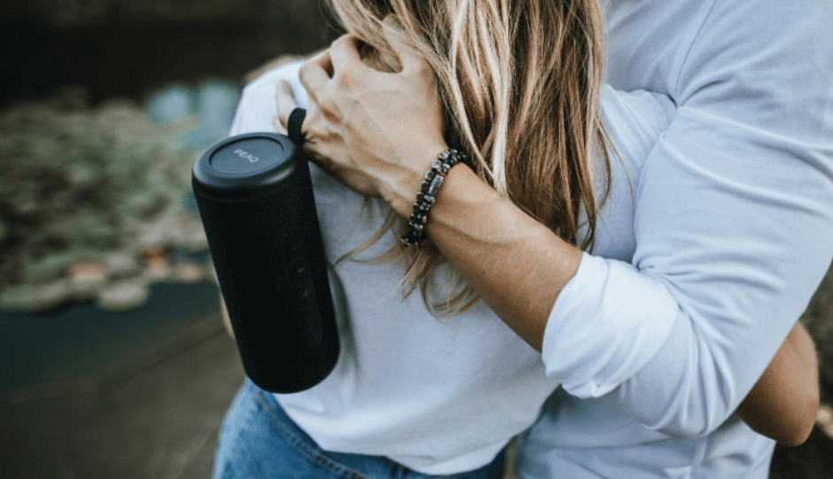 Un joven sostiene un altavoz bluetooth de la marca PEAQ, con su brazo rodea por los hombros a una mujer rubia, al aire libre, vista por detrás