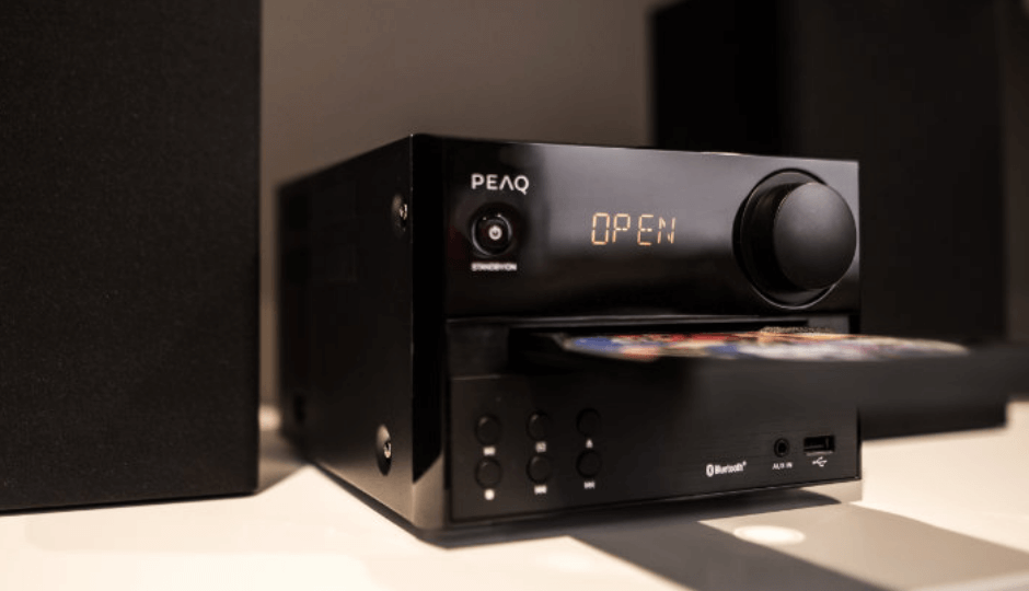 A PEAQ hi-fi system on a white table, next to black speaker boxes, CD being ejected from the device, close-up