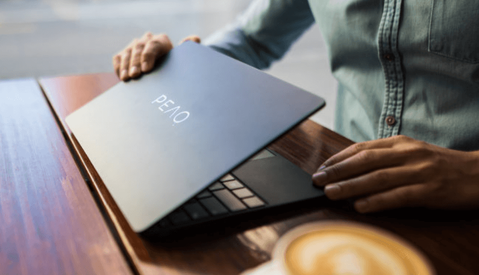 Jonge man opent een notebook van het merk PEAQ, op een houten tafel, daarnaast een volle kop koffie, in een café