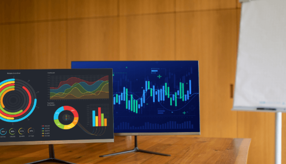 Due monitor a marchio PEAQ con grafici informativi, su un tavolo in legno in una sala conferenze, lavagna a fogli sullo sfondo