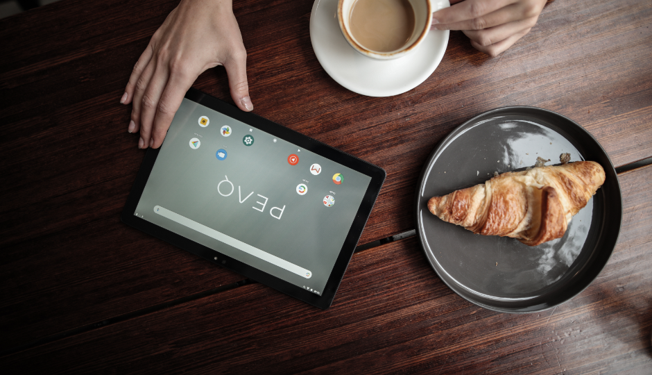 A PEAQ tablet on a kitchen table made of dark wood, next to a coffee cup and a croissant on a plate, and a hand reaching for the tablet