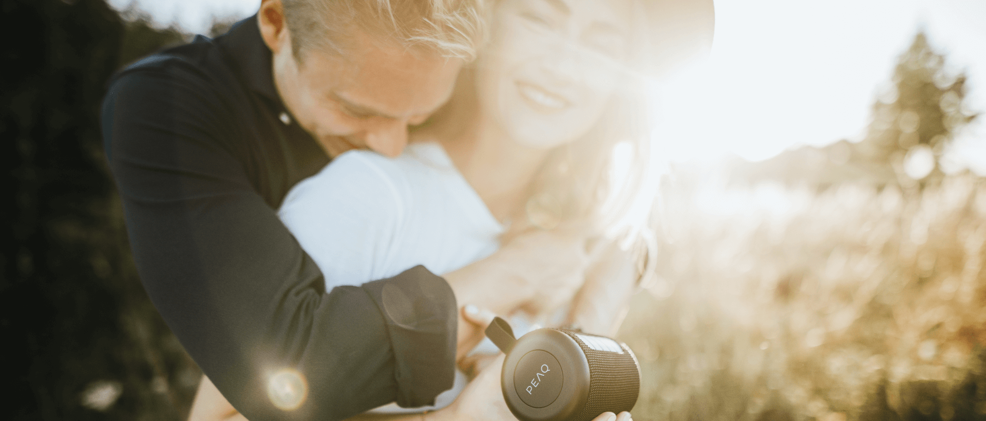 Een jonge vrouw houdt een bluetooth-luidspreker van het merk PEAQ in haar hand, een jonge man omarmt haar van achteren, ze lachen allebei, buiten, zon op de achtergrond