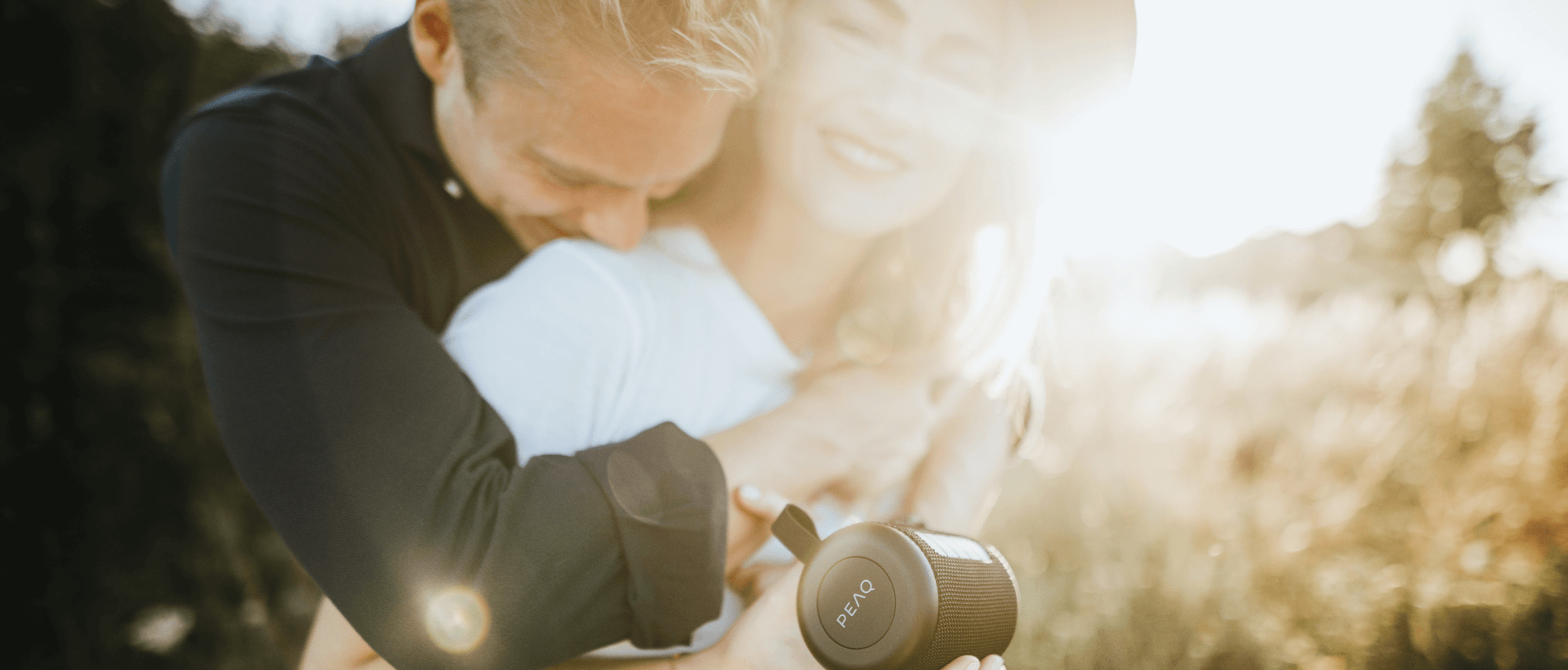 A young woman holding PEAQ Bluetooth speakers, a young man hugging her from behind, both laughing outdoors, sun in the background